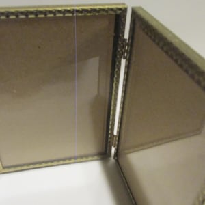 May include: A gold-toned metal picture frame with a decorative border. The frame is open and has two separate picture slots.