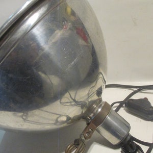 May include: A vintage silver metal spotlight with a black cord and a black switch. The spotlight has a large, round reflector and a small, round base.