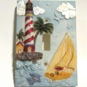 May include: A light switch cover with a 3D design of a lighthouse, palm tree, sailboat, and dolphins. The lighthouse is red and white striped with a yellow roof. The sailboat is yellow with a red sail. The dolphins are blue. The background is blue with white clouds.
