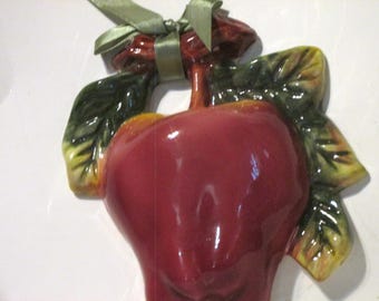May include: A ceramic apple ornament with a glossy red finish. The apple has green and yellow leaves and a green ribbon tied in a bow. A decorative piece for the kitchen or dining room.