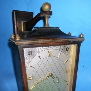 May include: A dark wood wall clock with a gold-toned face and Roman numeral hour markers. The clock has a decorative design with a sunburst pattern and a small moon and star motif. The clock is mounted on a wall with a decorative bracket.