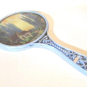 May include: A light blue hand mirror featuring an oval frame and an ornate, scrollwork handle. The mirror surface displays a printed landscape illustration of a cottage nestled among trees and greenery under a soft, warm-toned sky.