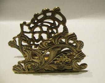 May include: A gold-toned metal napkin holder with an intricate, ornate design. The holder is made of a delicate, openwork pattern with swirls and floral motifs.