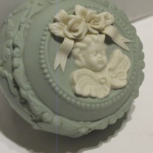 May include: A pale green ceramic trinket box with a raised design of an angel with wings and flowers. The angel has a white face and is wearing a white ribbon.