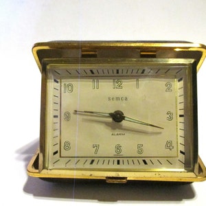 May include: A gold-toned, square-shaped alarm clock with a white face. The clock has black numbers and hands, and the word "semca" is printed in black on the face. The clock is in a closed position, with the lid closed.