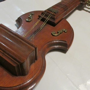 May include: A wooden wall-mounted violin-shaped shelf with a small shelf on the side. The shelf is decorated with gold accents and has a stringed instrument design.