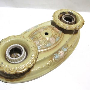 May include: Vintage two-light ceiling fixture with a cream-colored finish. The fixture features two bulb sockets and a decorative floral design with pink and blue accents. The fixture is made of metal and has a distressed appearance.