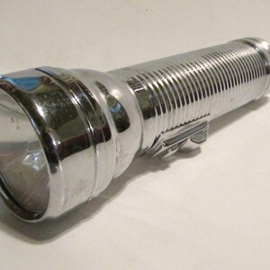 May include: A cylindrical flashlight with a polished chrome finish. The body features a ribbed texture for grip, and a rectangular sliding switch is located on the side. The head of the flashlight is smooth and rounded, housing a clear lens.