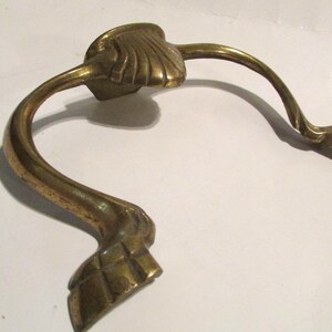 May include: A brass drawer pull featuring a central shell-shaped mounting plate and two curved arms that extend outward, terminating in detailed, foot-like ends. The metal has a polished, golden-toned finish with visible signs of age and wear.