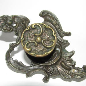 May include: A brass drawer pull featuring a circular knob centered on an ornate, asymmetrical backplate with intricate scrollwork and leaf-like detailing. The metal has a darkened, aged patina with highlights on the raised surfaces.