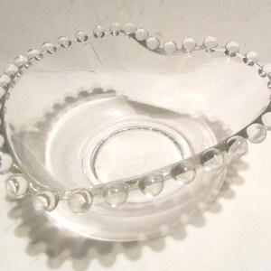May include: A clear glass bowl featuring a heart-shaped rim decorated with a series of small, spherical glass beads along the entire edge. The bowl has a smooth, transparent interior and a circular base, providing a simple and functional design.