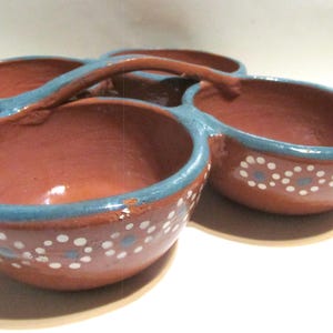 May include: A terracotta-colored serving dish with four connected bowls and a handle. Each bowl has a blue rim and is decorated with white and blue floral patterns. The dish is likely handmade and suitable for serving snacks or dips.