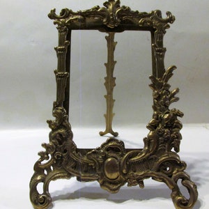 May include: A gold-toned metal picture frame with ornate details and a decorative base. The frame is designed to stand upright and has a rectangular opening for a picture.