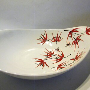 May include: White ceramic serving bowl with a red and black floral design. The bowl is oval shaped and has a slight curve on one side.
