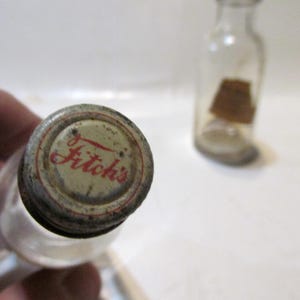 May include: Close-up of a vintage glass bottle with a metal cap. The cap is cream-colored with the red text "Fitch's". A second, similar bottle is in the background. The bottles appear to be old and possibly antique.