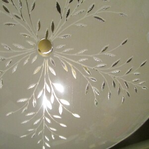 May include: A frosted glass ceiling light fixture with a gold center and a leaf design. The leaves are etched in silver and gold, radiating outwards from the center. The fixture is round and has a soft, diffused glow.
