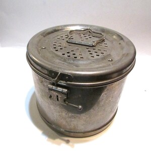 May include: A vintage, cylindrical metal container with a perforated lid and a hinged clasp. The container has a handle on the lid and a latch on the side. The metal has a worn, aged appearance.