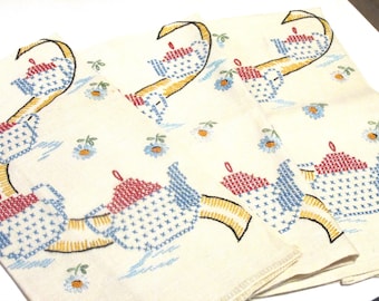May include: Cream-colored embroidered napkins with a teapot design. The teapots are red and blue, with yellow handles and spouts. Small blue and white flowers are scattered across the fabric.