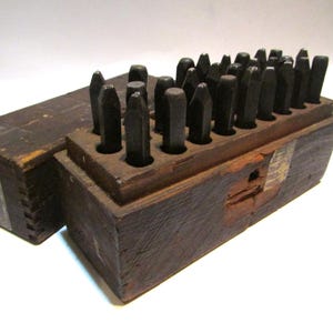 May include: A vintage wooden box with a set of metal letter stamps. The stamps are arranged in a wooden holder, with the box lid open beside it. The wood has a dark, aged appearance, suggesting it is an antique tool.