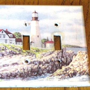 May include: A white ceramic light switch cover with a colorful image of a lighthouse on a rocky coastline. The switch cover has two toggle switches.