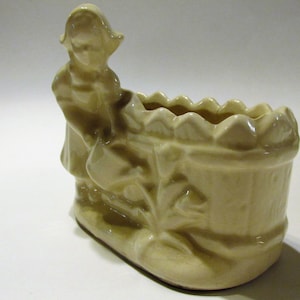 May include: A cream-colored ceramic planter shaped like a girl pushing a wheelbarrow. The planter has a scalloped edge and is decorated with flowers.