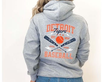 May include: Grey hooded sweatshirt with a vintage-style graphic of the Detroit Tigers baseball team. The graphic features a baseball, bats, and the text "DETROIT Tigers EST. 1894 CENTRAL DIVISION BASEBALL".