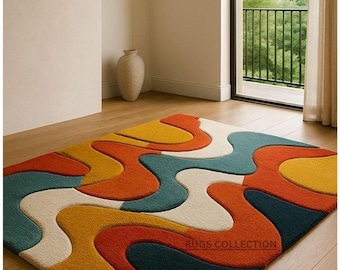May include: A colorful area rug with an abstract design. The rug features flowing, organic shapes in shades of orange, yellow, teal, white, and navy blue. The rug is placed on a wooden floor in a room with a window and a neutral-colored wall.