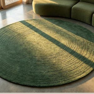 May include: A round, olive green rug with a concentric design. The rug is placed on a concrete floor, with a green sofa in the background. The rug's diameter is approximately 7 feet.