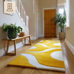 May include: A hallway runner rug with a yellow and white abstract design. The rug is rectangular and features a flowing pattern of yellow and white. It's laid on a wooden floor, with a bench and plants visible. The walls are painted in neutral colours. The rug's dimensions are approximately 200cm x 80cm.