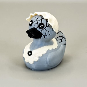 May include: A whimsical, light blue duck figurine with a cracked eggshell design on its head and body. The duck has a black beak and eyes, and a white ruffled collar. The figurine is a unique decorative item.