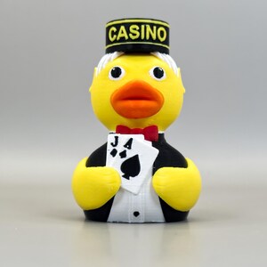 May include: A yellow rubber ducky dressed as a casino dealer, wearing a black hat with "CASINO" in yellow letters. The duck holds playing cards and a red bow tie. The duck has a white shirt and black tuxedo.