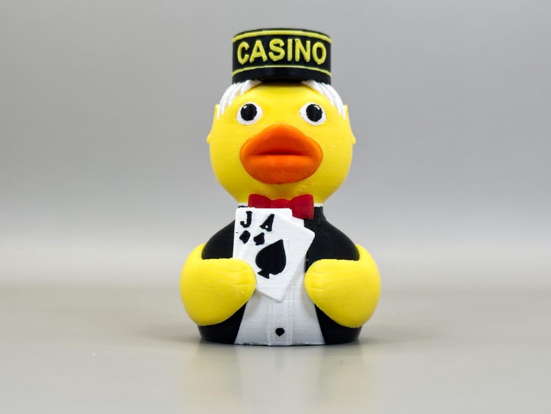May include: A yellow rubber ducky dressed as a casino dealer, wearing a black hat with "CASINO" in yellow letters. The duck holds playing cards and a red bow tie. The duck has a white shirt and black tuxedo.