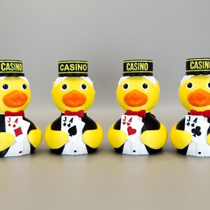May include: Four yellow rubber duck figurines dressed as casino workers. Each duck wears a black and gold hat with "CASINO" printed on it, a red bow tie, and a tuxedo. Each duck holds a playing card.