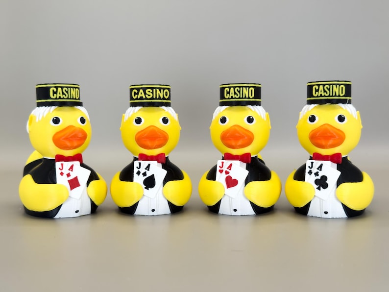 May include: Four yellow rubber duck figurines dressed as casino workers. Each duck wears a black and gold hat with "CASINO" printed on it, a red bow tie, and a tuxedo. Each duck holds a playing card.