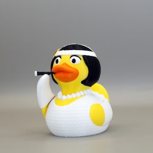 May include: A novelty rubber ducky figurine, styled as a flapper. The yellow duck wears a black bob wig, white headband, and a pearl necklace. It holds a cigarette holder in its beak and is dressed in a white dress with yellow accents.