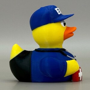 May include: A yellow rubber duck wearing a blue baseball cap with a white "E" on it, a blue uniform, and a stethoscope. The duck has an orange beak and is holding a red first aid kit.