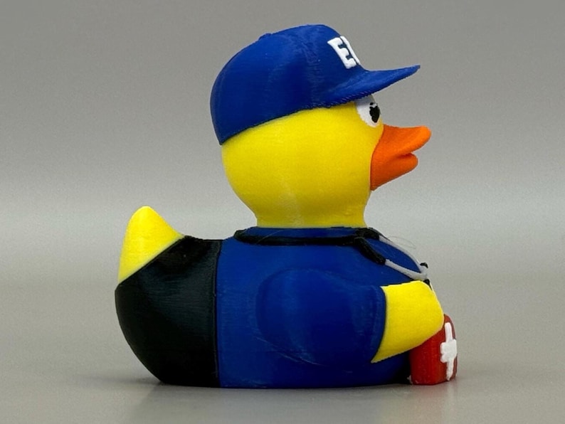 May include: A yellow rubber duck wearing a blue baseball cap with a white "E" on it, a blue uniform, and a stethoscope. The duck has an orange beak and is holding a red first aid kit.