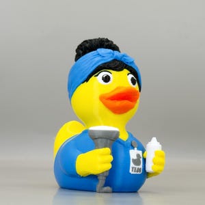 May include: A yellow rubber duck figurine dressed as a medical technician. The duck has black hair in a bun with a blue headband, wears a blue scrub top, and holds a gray ultrasound probe and a white bottle. An ID badge reads TECH with a duck icon.