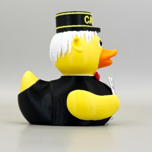 May include: A yellow rubber duck wearing a black suit, a red bow tie, and a black hat with the word "CA" in gold lettering. The duck has white hair and an orange beak. It is holding a small white object.