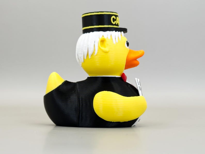 May include: A yellow rubber duck wearing a black suit, a red bow tie, and a black hat with the word "CA" in gold lettering. The duck has white hair and an orange beak. It is holding a small white object.