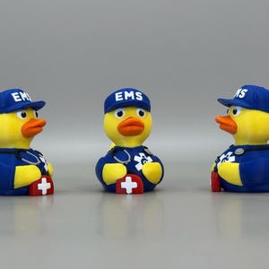 May include: Three rubber duck figurines dressed as emergency medical service (EMS) workers. Each duck wears a blue cap with "EMS" on it, a blue uniform, and a stethoscope. The ducks have yellow bodies, orange beaks, and red crosses.