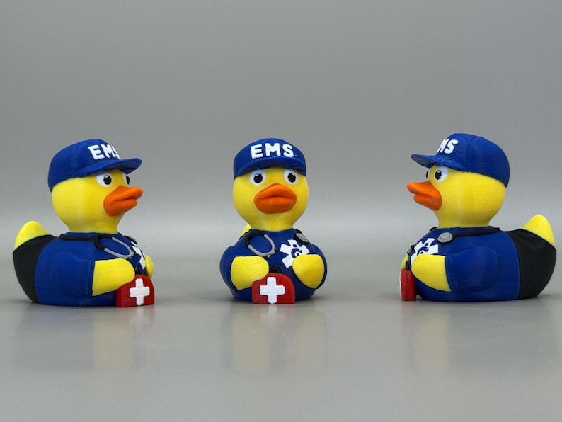 May include: Three rubber duck figurines dressed as emergency medical service (EMS) workers. Each duck wears a blue cap with "EMS" on it, a blue uniform, and a stethoscope. The ducks have yellow bodies, orange beaks, and red crosses.