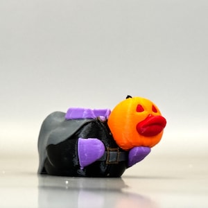 May include: A 3D-printed Halloween-themed figurine. It features an orange pumpkin head with red lips and eye cutouts, a black body, and purple accents. The figurine has a belt and a cape-like structure, creating a whimsical character.