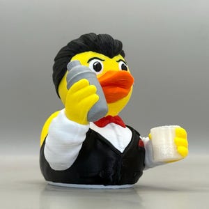 May include: A yellow rubber duck figurine dressed as a bartender, featuring black hair, a black vest, a white shirt, and a red bowtie. The duck holds a grey cocktail shaker in one wing and a small, translucent white glass in the other wing.