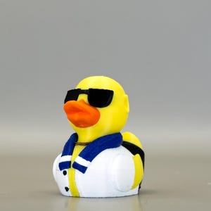 May include: A yellow rubber duck wearing black sunglasses, a blue and white scarf, and a white jacket with black buttons. The duck has an orange beak and is a fun, novelty item.