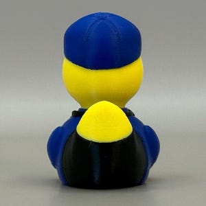 May include: A 3D-printed figurine of a character with a yellow head and a blue hat and uniform. The figure has a black lower body and a yellow backpack. The background is a solid, light gray color.