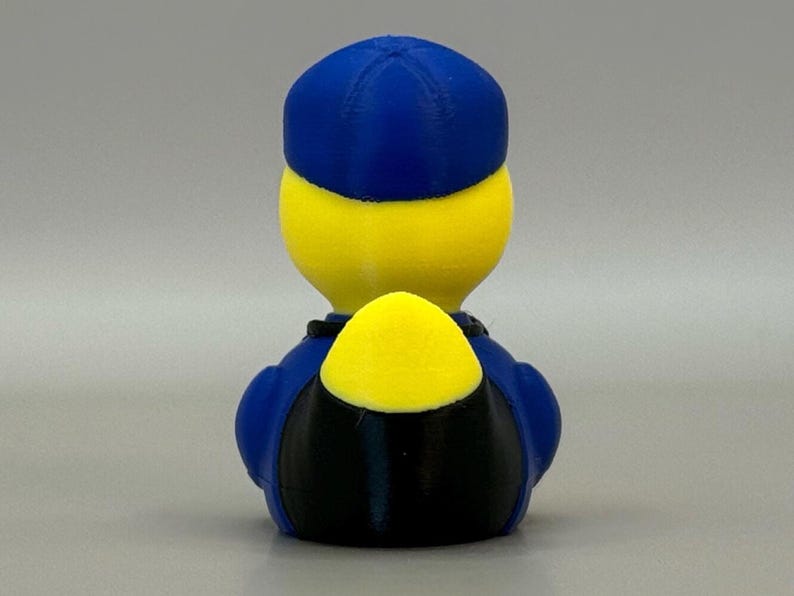 May include: A 3D-printed figurine of a character with a yellow head and a blue hat and uniform. The figure has a black lower body and a yellow backpack. The background is a solid, light gray color.