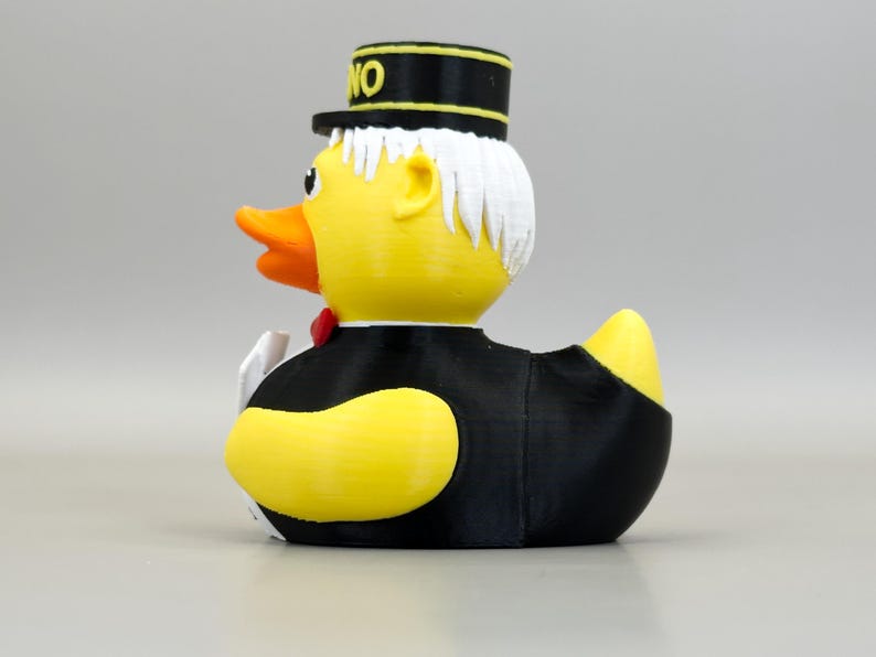 May include: A yellow rubber duck wearing a black tuxedo, a red bow tie, and a black hat with the word "NO" on it. The duck has white hair and an orange beak. A fun novelty item.