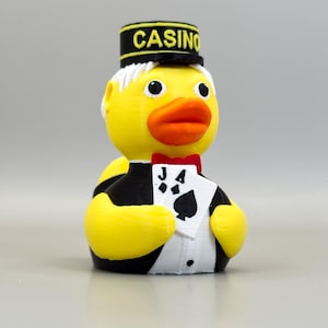 May include: A yellow rubber ducky dressed as a casino worker, wearing a black hat with "CASINO" in yellow letters. The duck has a red bow tie and is holding a playing card with the Jack of Spades.
