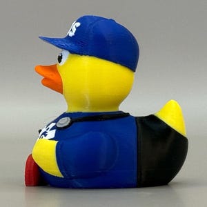 May include: A yellow rubber duck wearing a blue baseball cap with white lettering and a blue uniform. The duck has an orange beak and red feet. The duck is a novelty item.